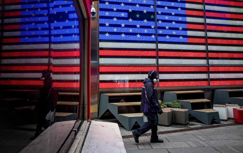 △一名美国快递员工佩戴着口罩走过时代广场（图片来源：彭博社）